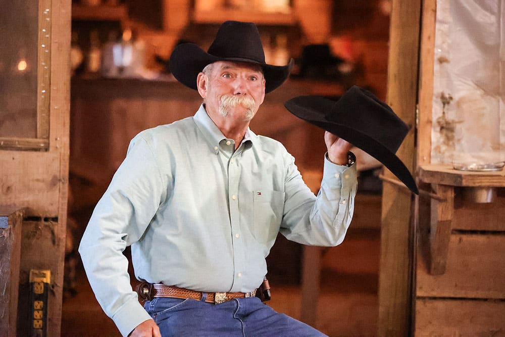 A cowboy with a gray mustache, wearing a black cowboy hat and holding a black cowboy hat.